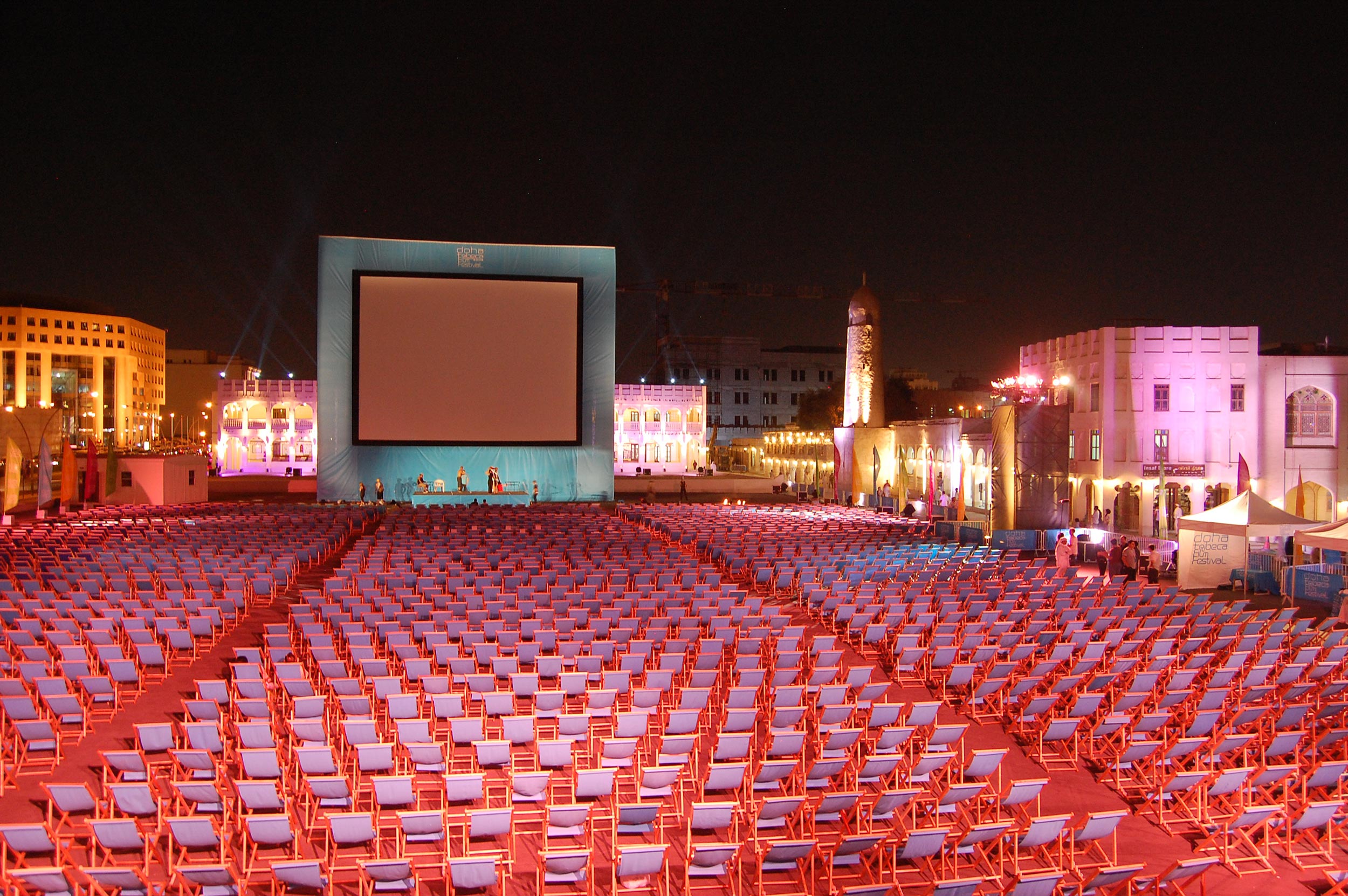 Outdoor cinema setup at night with hundreds of empty chairs facing a large screen in a lit plaza