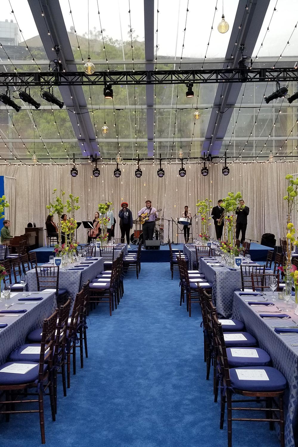 Elegant event setup with banquet tables, floral centerpieces, and a live band performing on stage under a clear canopy roof