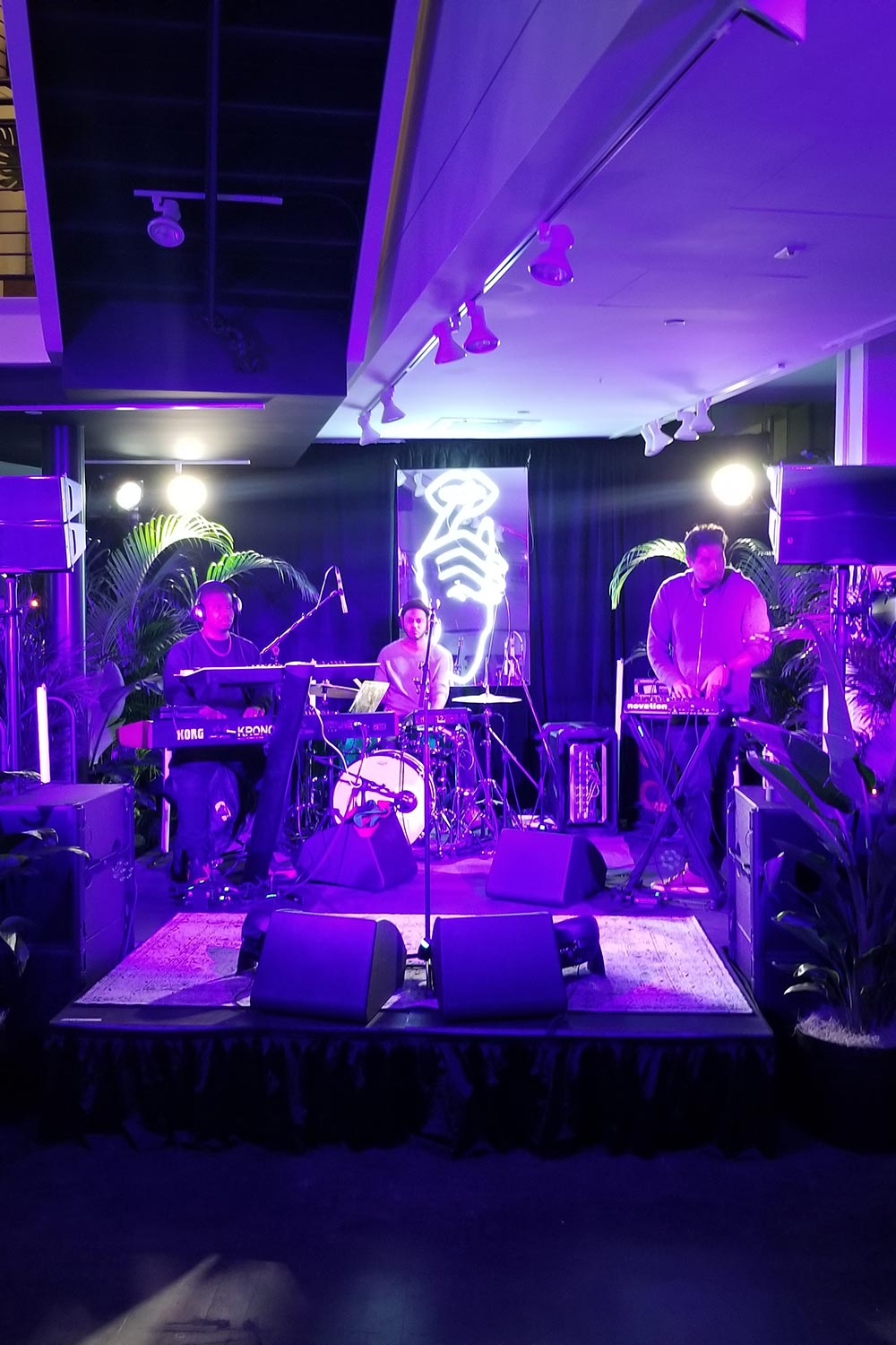 Live band performing on stage with purple lighting and a neon microphone sign in the background