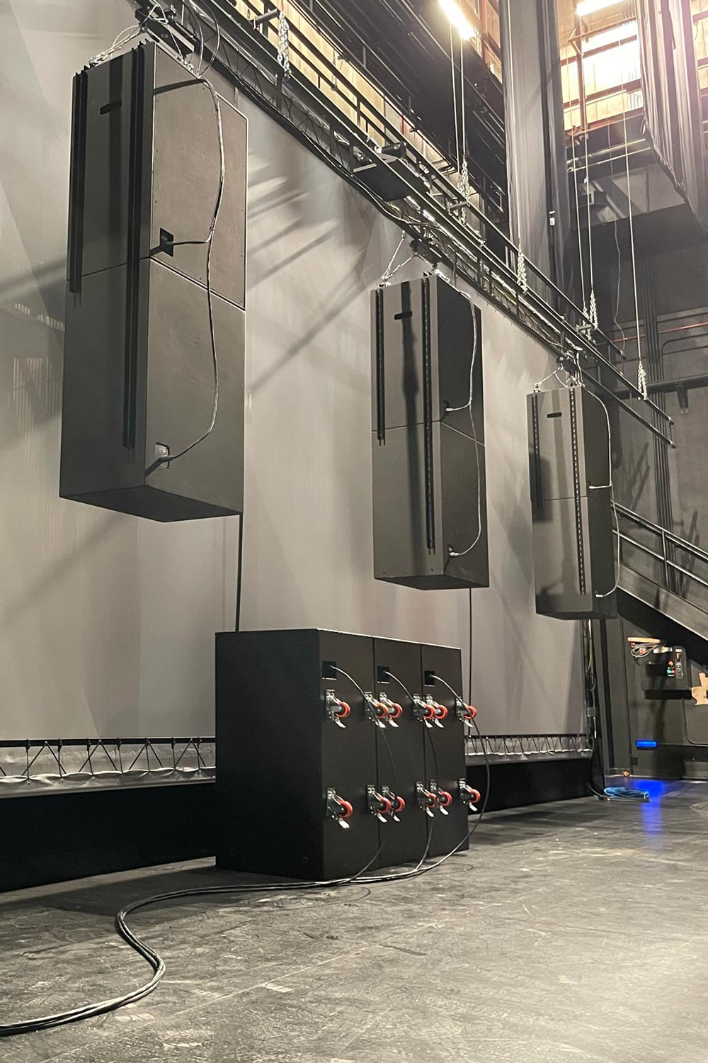 Large black speakers suspended on stage with subwoofers on floor connected by cables
