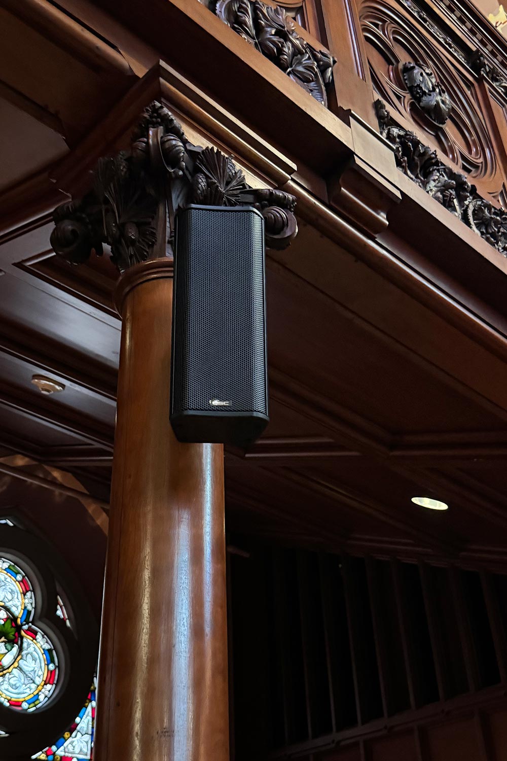 Black speaker mounted on a wooden column with ornate carvings in a historic building interior