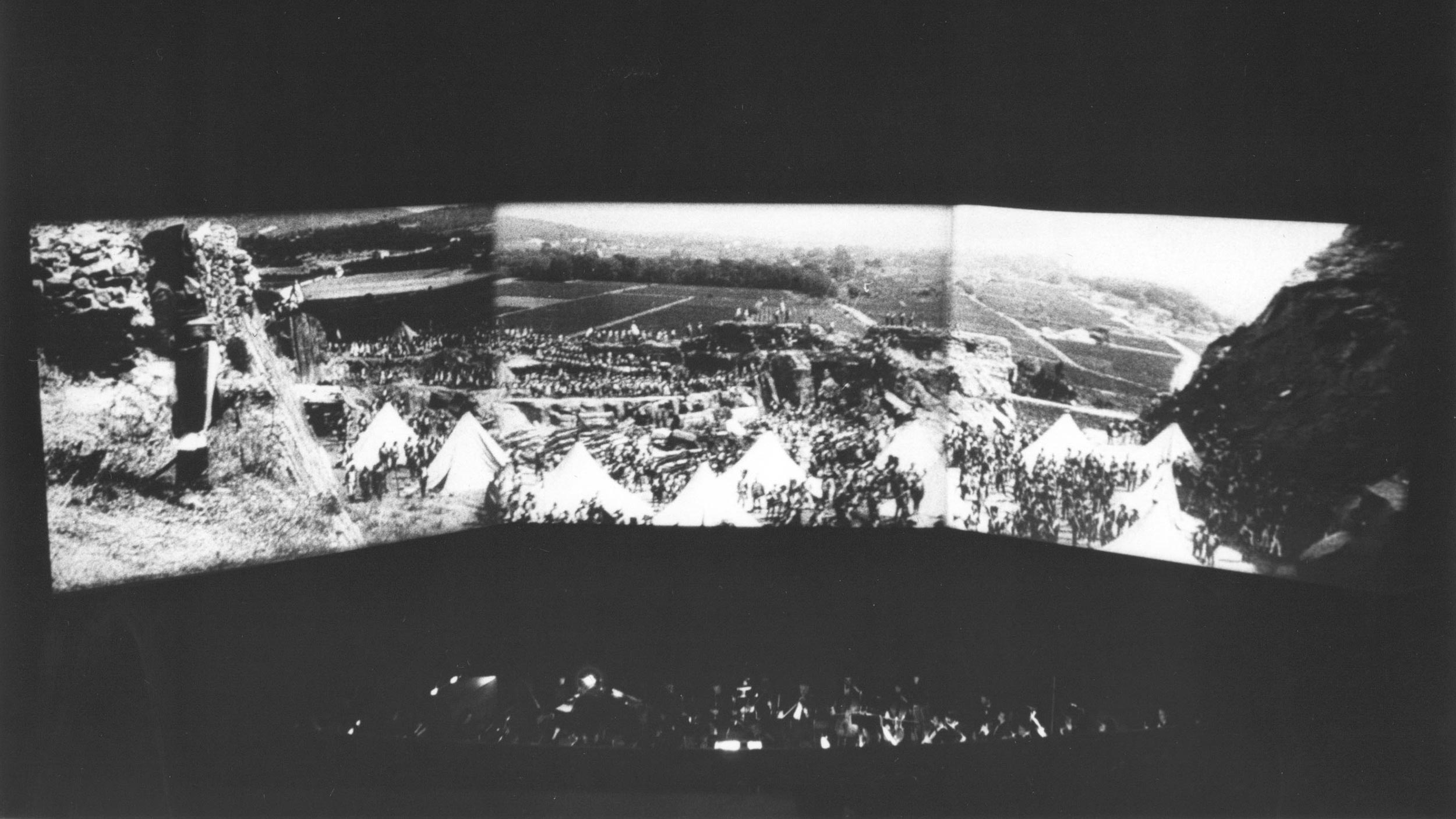 Wide three-panel projection of a historical battlefield scene with orchestra below