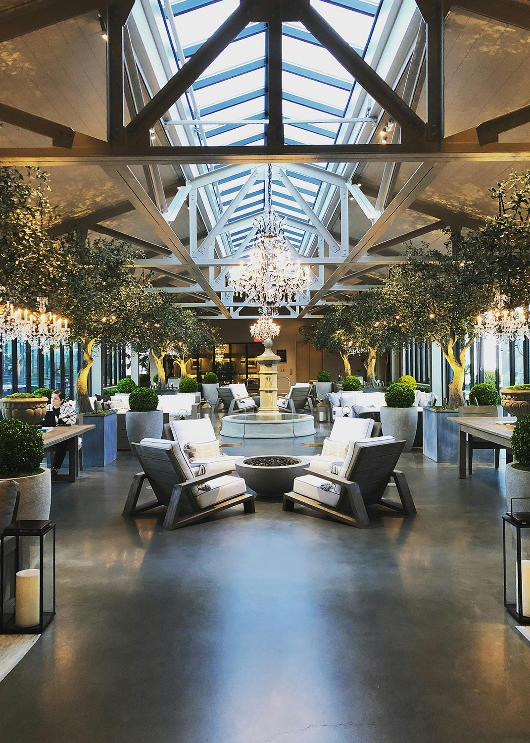 A luxurious indoor event space with chandeliers, greenery, and cozy seating areas under a skylight.