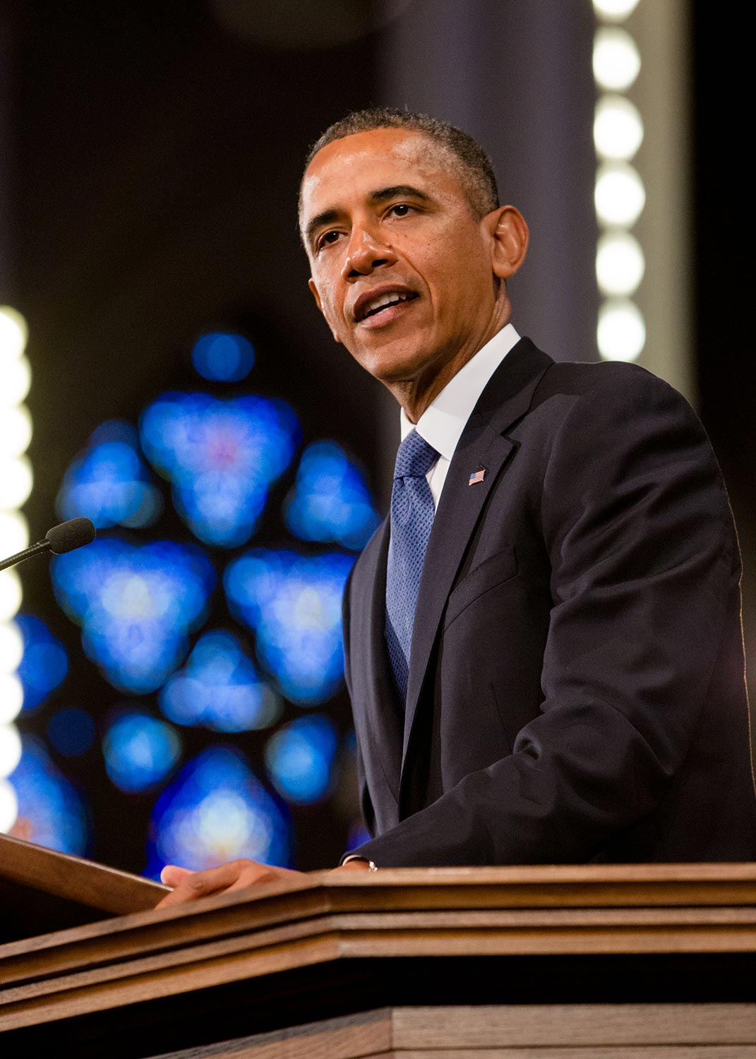 A speaker in a suit delivering a speech at a podium with a blurred stained glass window in the background.