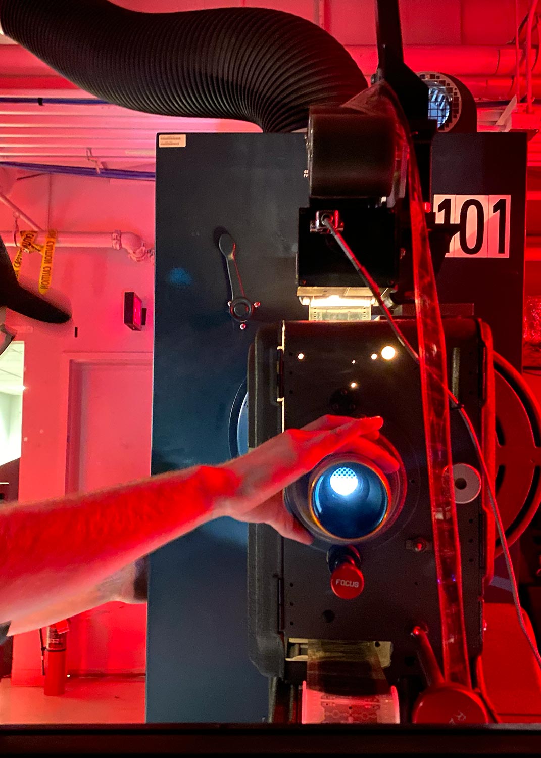 A person adjusting the lens of a film projector in a dimly lit projection room with red lighting.