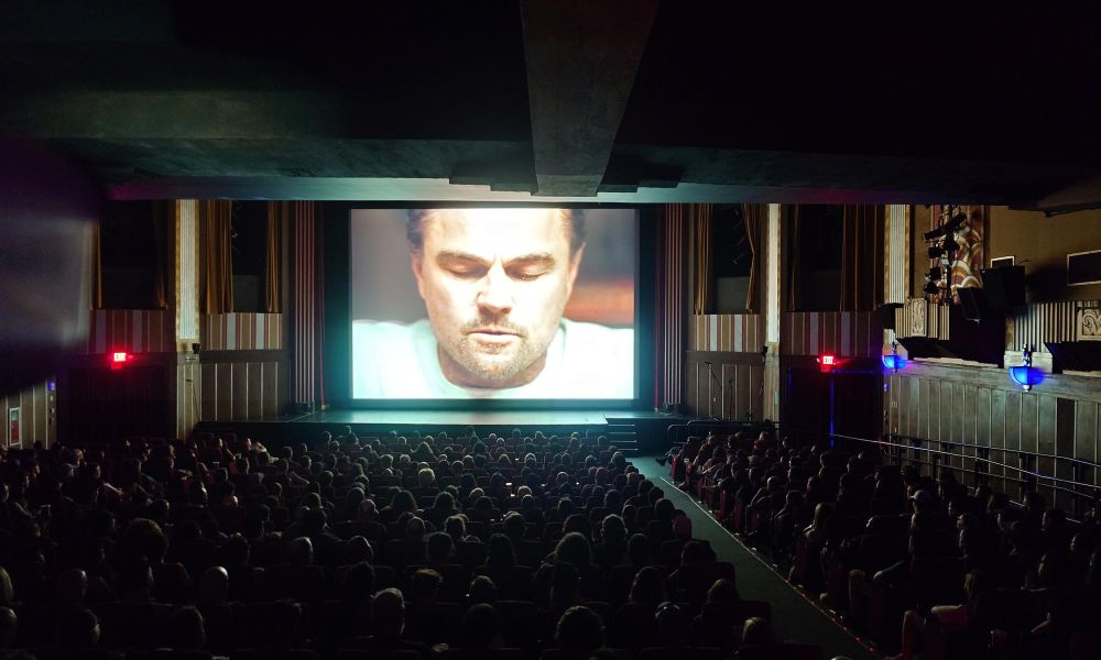 Audience in a dark theater watching a close-up of a man on a large movie screen