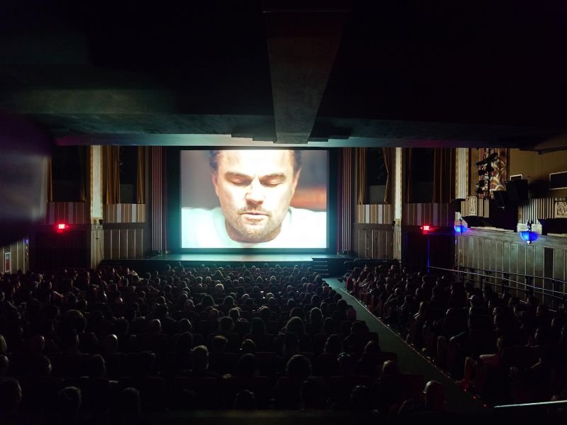 Audience in a dark theater watching a close-up of a man on a large movie screen