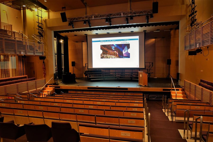 Auditorium with tiered wooden seating, central stage featuring a large projection screen and podium