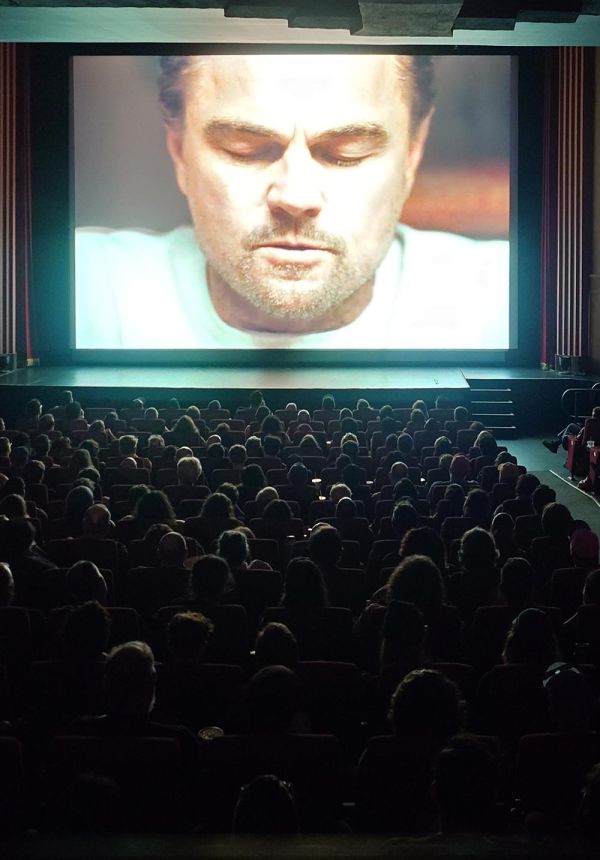 Crowded cinema seats facing a large illuminated screen with a man's face