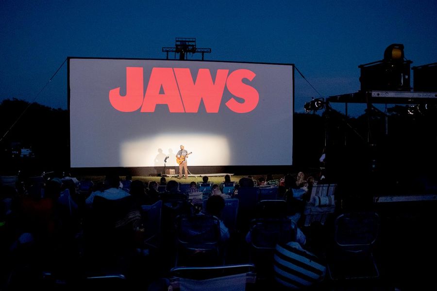 Evening outdoor movie event showing the JAWS title on screen with a live musician performing on stage in front