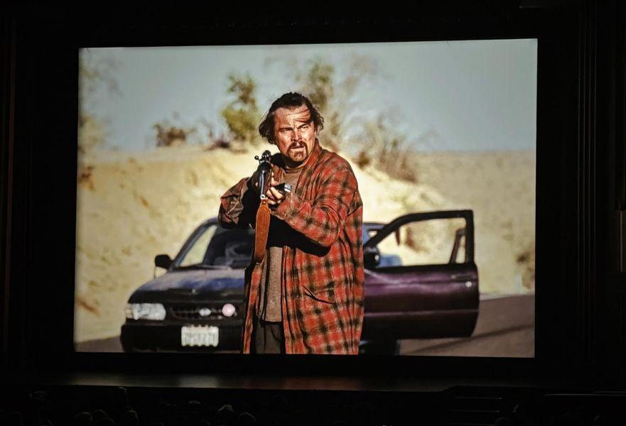 Movie screen showing a man in a plaid jacket holding a rifle next to a car