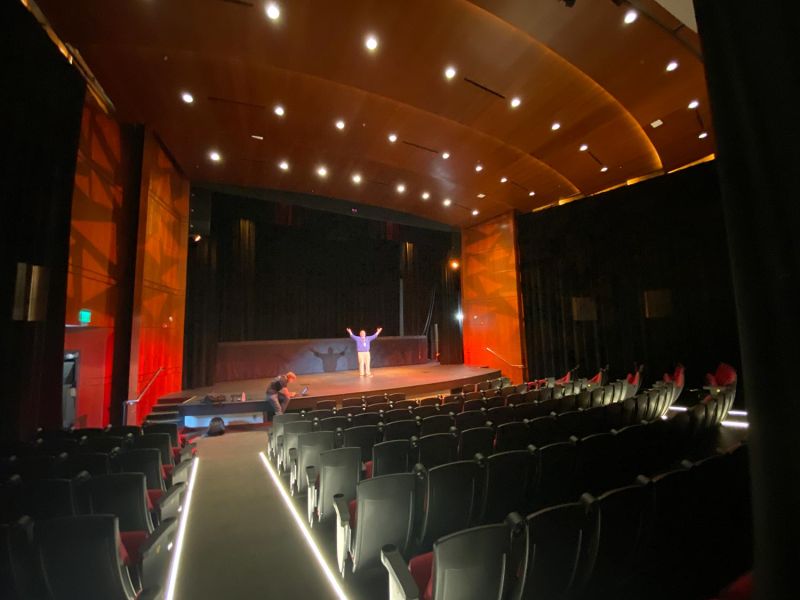 Wide view of an empty theater stage with a person standing in the center and arms raised